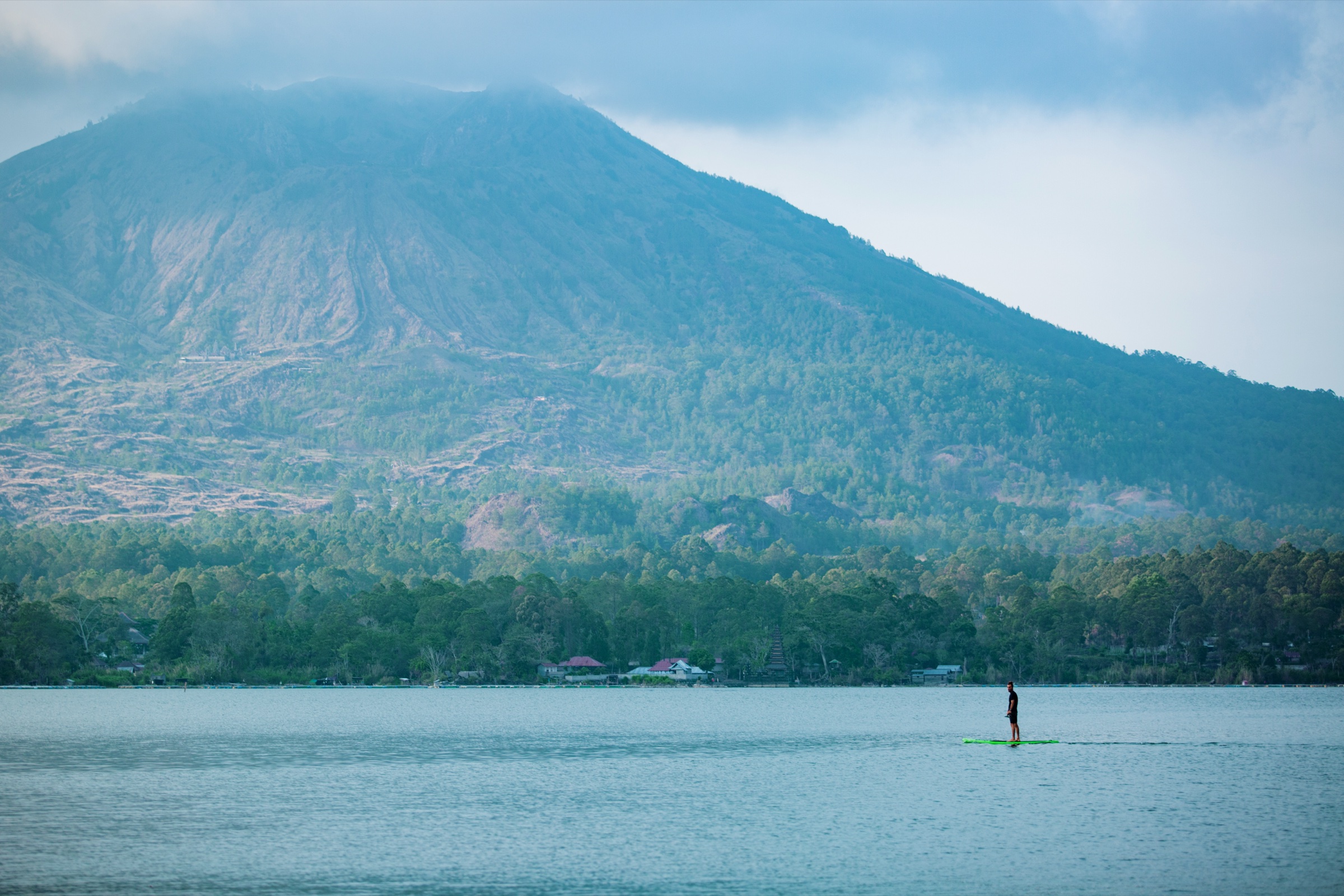 A distant paddleboarder on a calm lake beneath a forested, cloud-topped mountain.