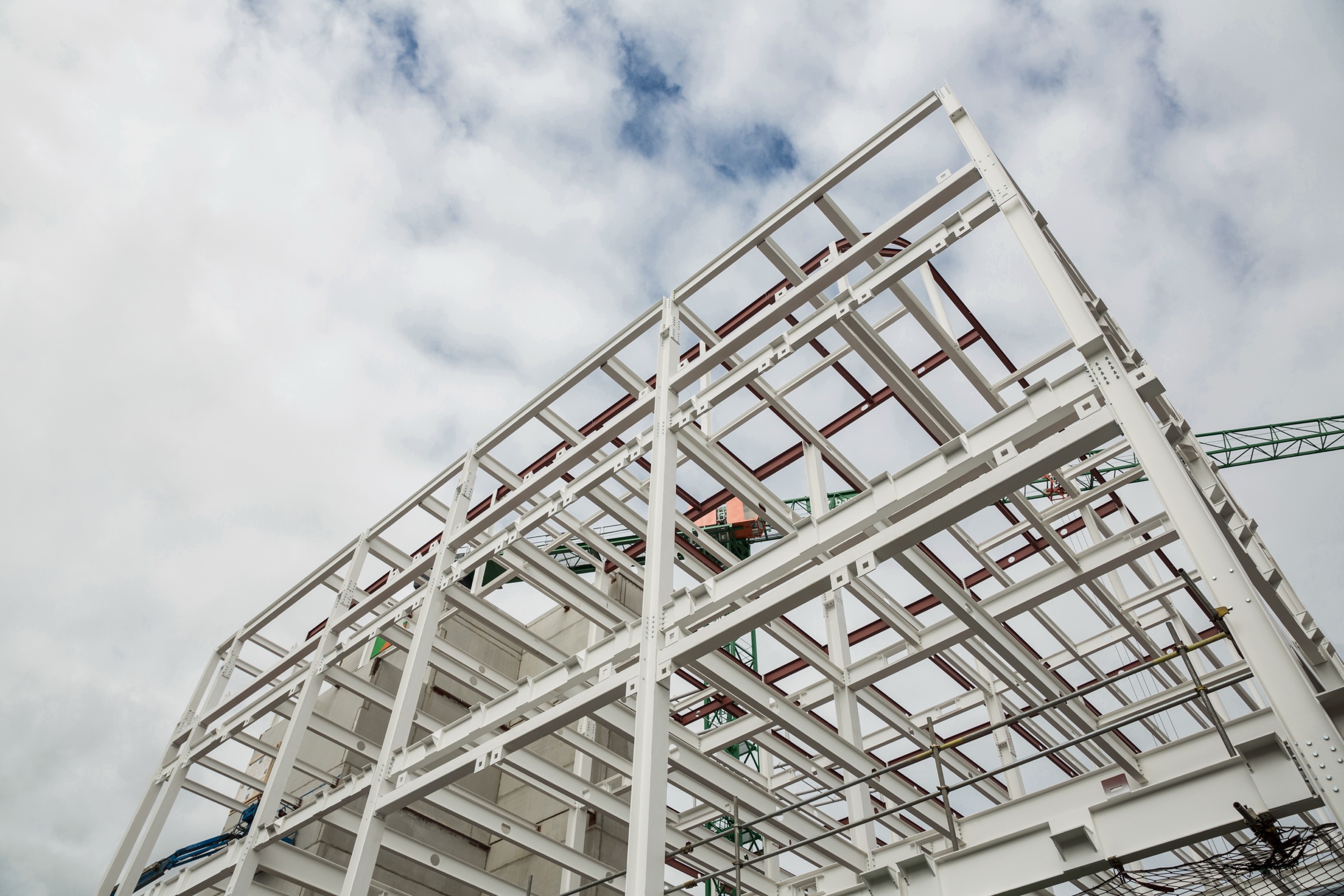 A white steel building frame under construction seen from below against a cloudy sky.