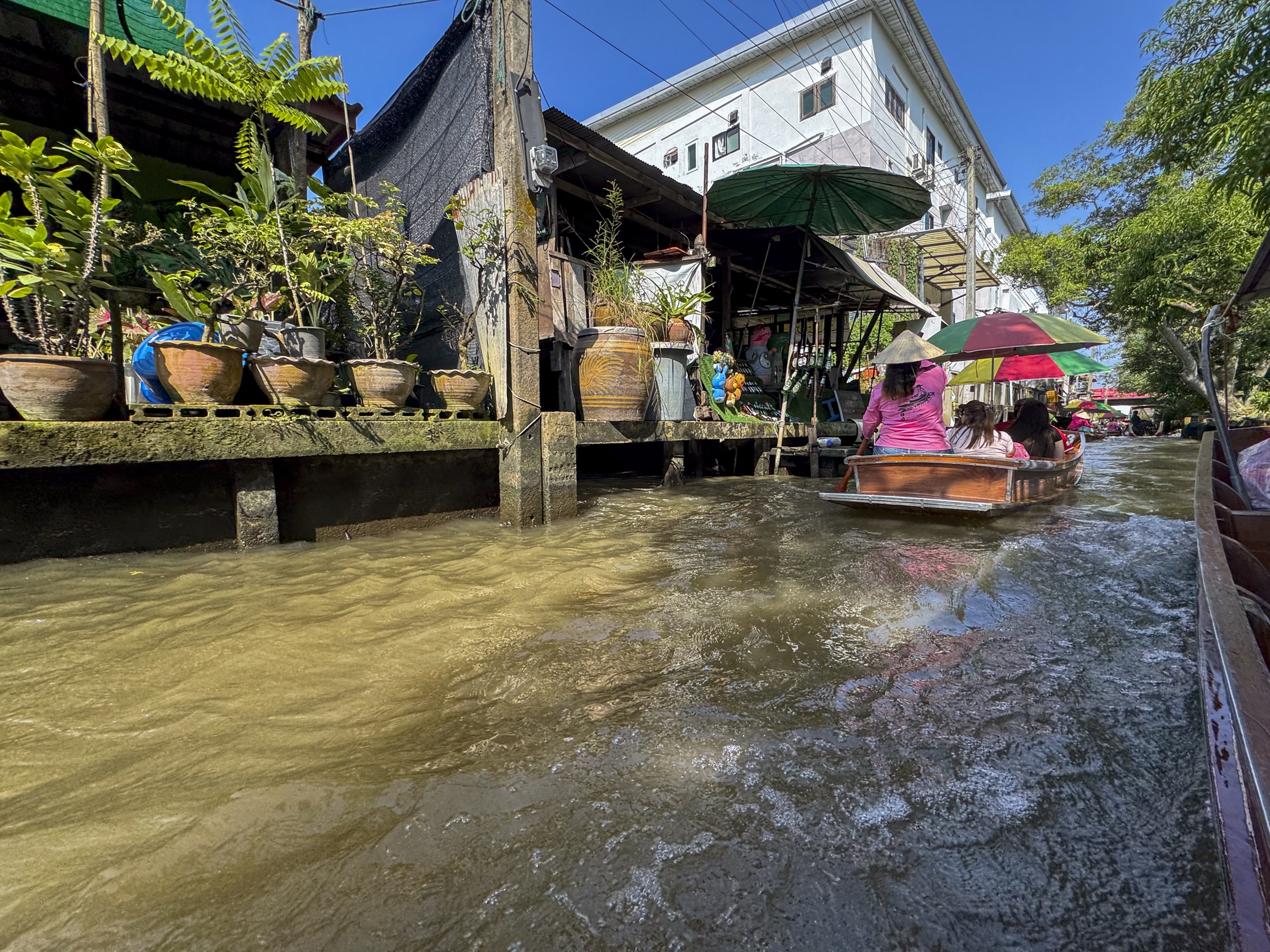 Brown canal water rises near a wooden platform of potted plants, with a boat and umbrellas in the distance.