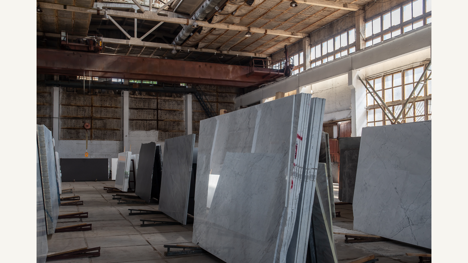 Illustration of upright polished stone slabs arranged in rows inside a large industrial warehouse, with reflective surfaces, vertical supports and open floor space visible.