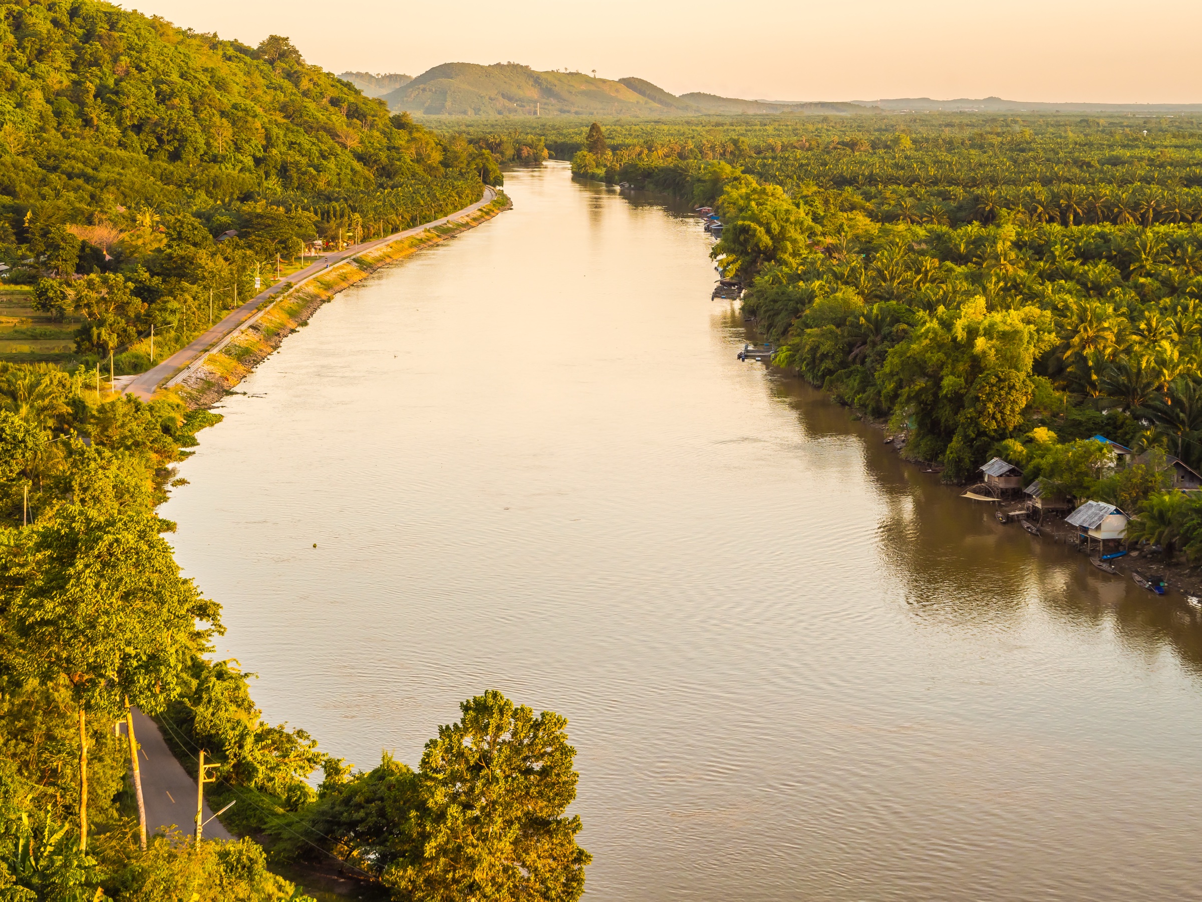 Aerial view of a wide river at sunset with green forest on both banks and a narrow road beside the water.